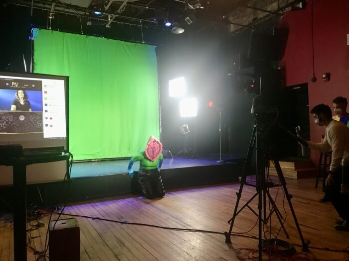 A performer wears an elaborate, textured pink vulva head piece and mask. They kneel in front of a green screen. On the edges of the image we see production screens, lights, and crew members.