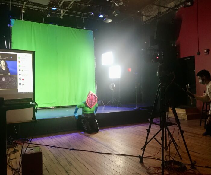 A performer wears an elaborate, textured pink vulva head piece and mask. They kneel in front of a green screen. On the edges of the image we see production screens, lights, and crew members.