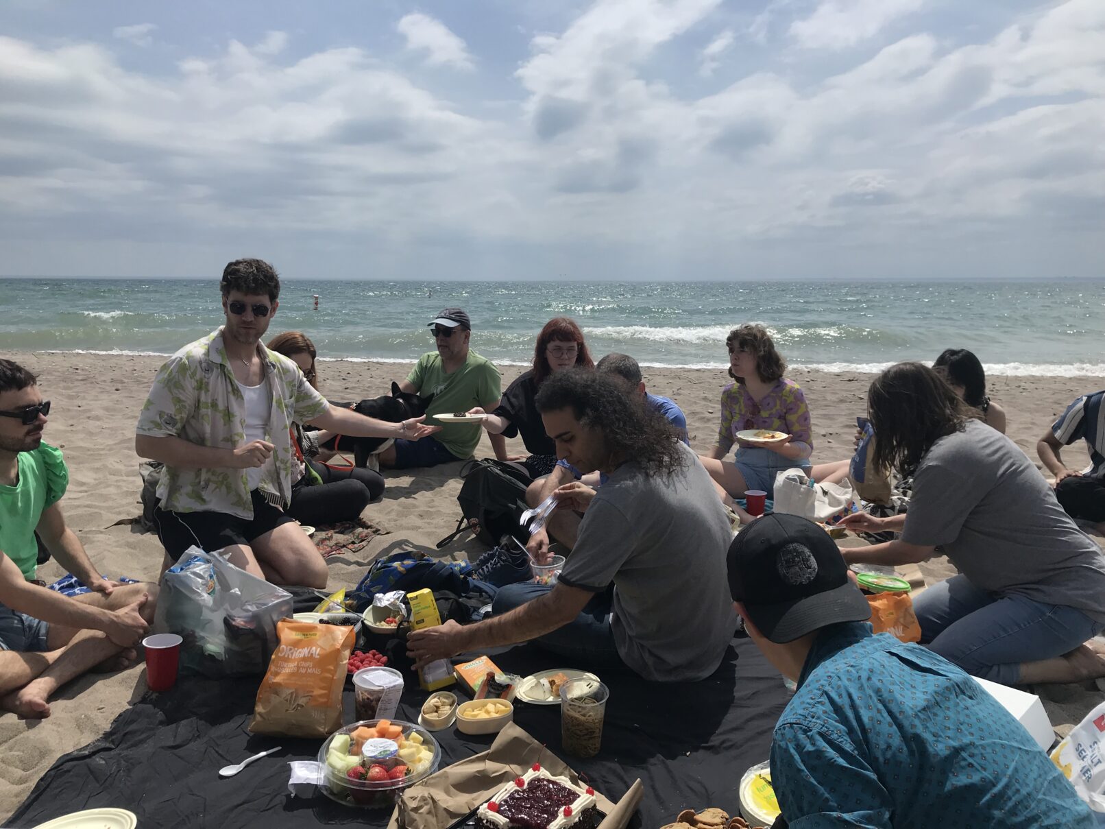 A group of people picnic on the beach. The sky is blue and grey and the water is a dark and moody aqua.