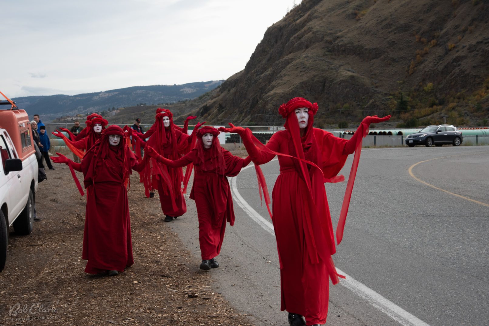 Performers dressed in bright red garments walk along a mountain highway. Their arms are extended out; red gloved palms to the sky. This is a photo of the Red Rebel Brigade in front of pipes for the TransMountain Pipeline near Kamloops, British Columbia, Canada.