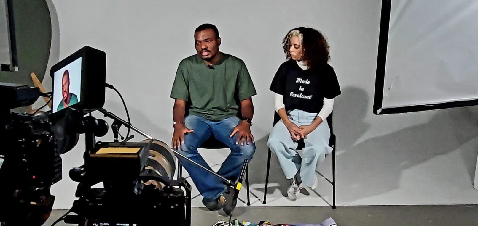 A photo of Pablo Carvalho and Tais Almeida. They are being interviewed. Pablo is a young black man wearing jeans and t-shirt. aTais is a young mixed race black woman. She also wears jeans and a tshirt. We see the camera and lighting screens in the frame of the photo.
