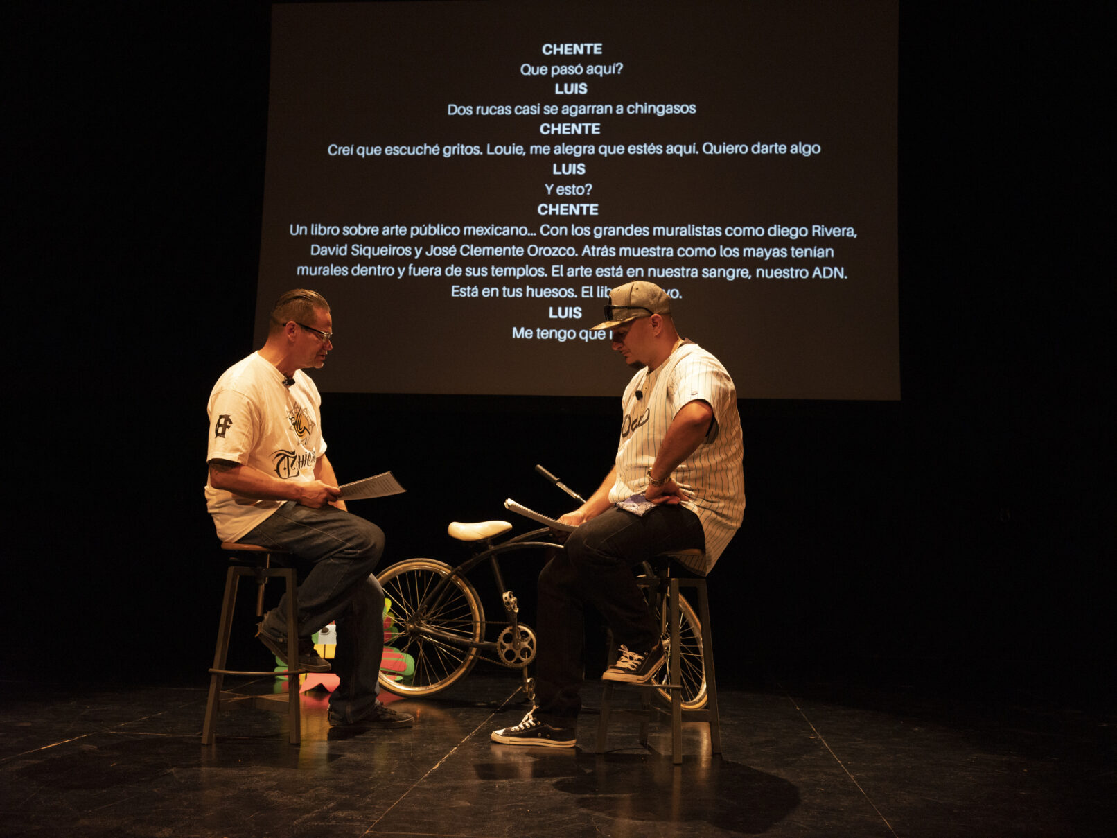 Two men on stage read from a script; text is projected behind them. Onstage is a bicycle.