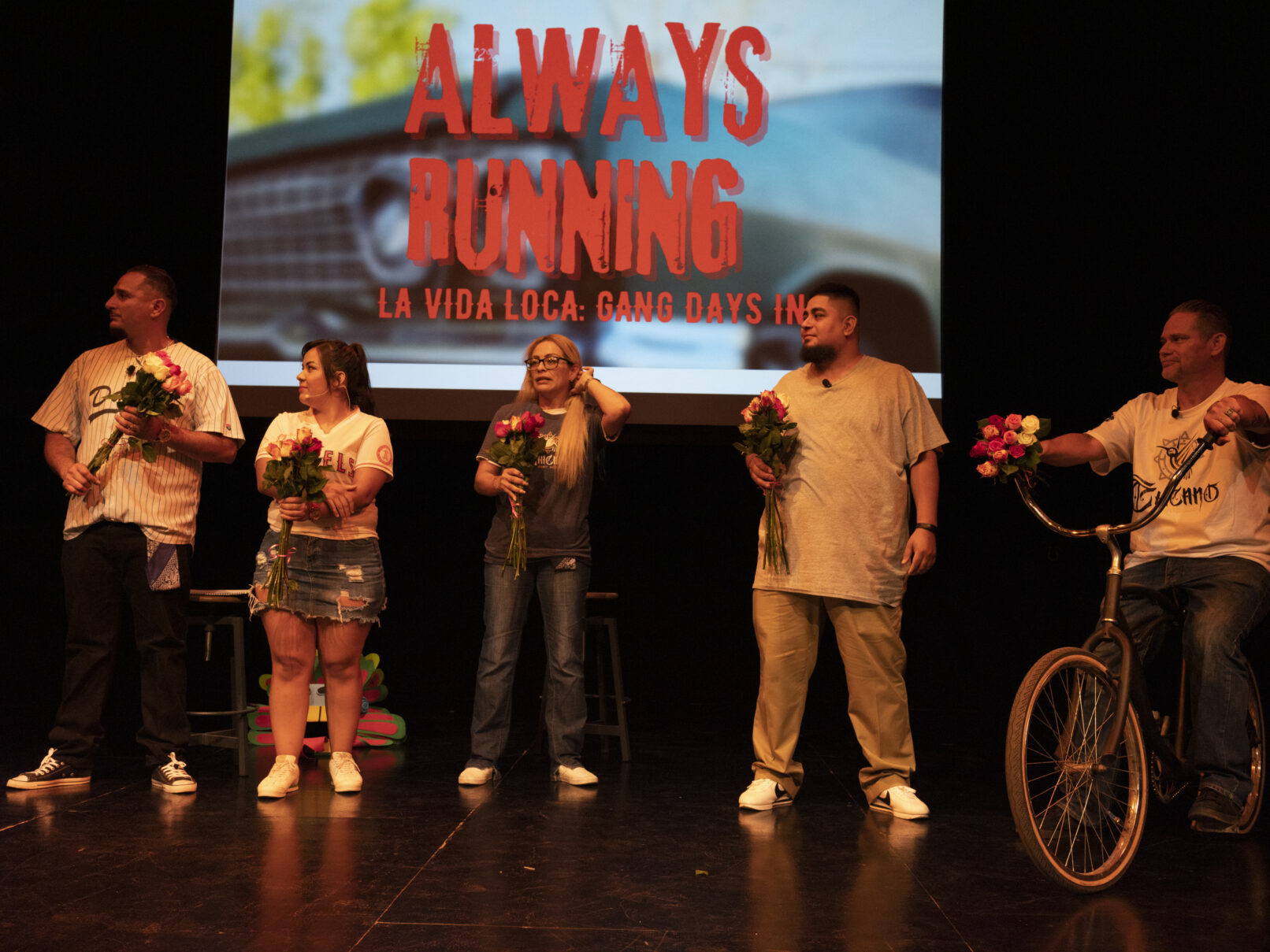 The cast of Always Running stands on stage. Behind them is projected the title of the play ontop of a blue car. The text reads: Always Running: La Vida Loca