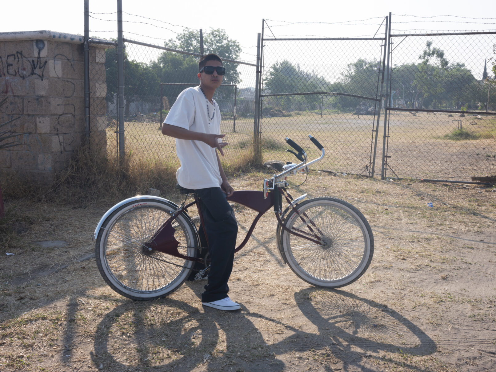 A young man poses on his bike in front of a chain link fence. He makes a gang sign with his right hand.