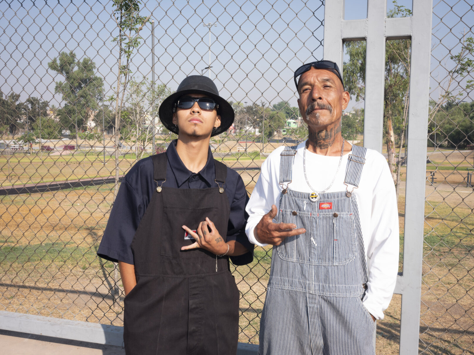 Two men pose for the camera in from of a chain link fence. They make gang signs with their hands.