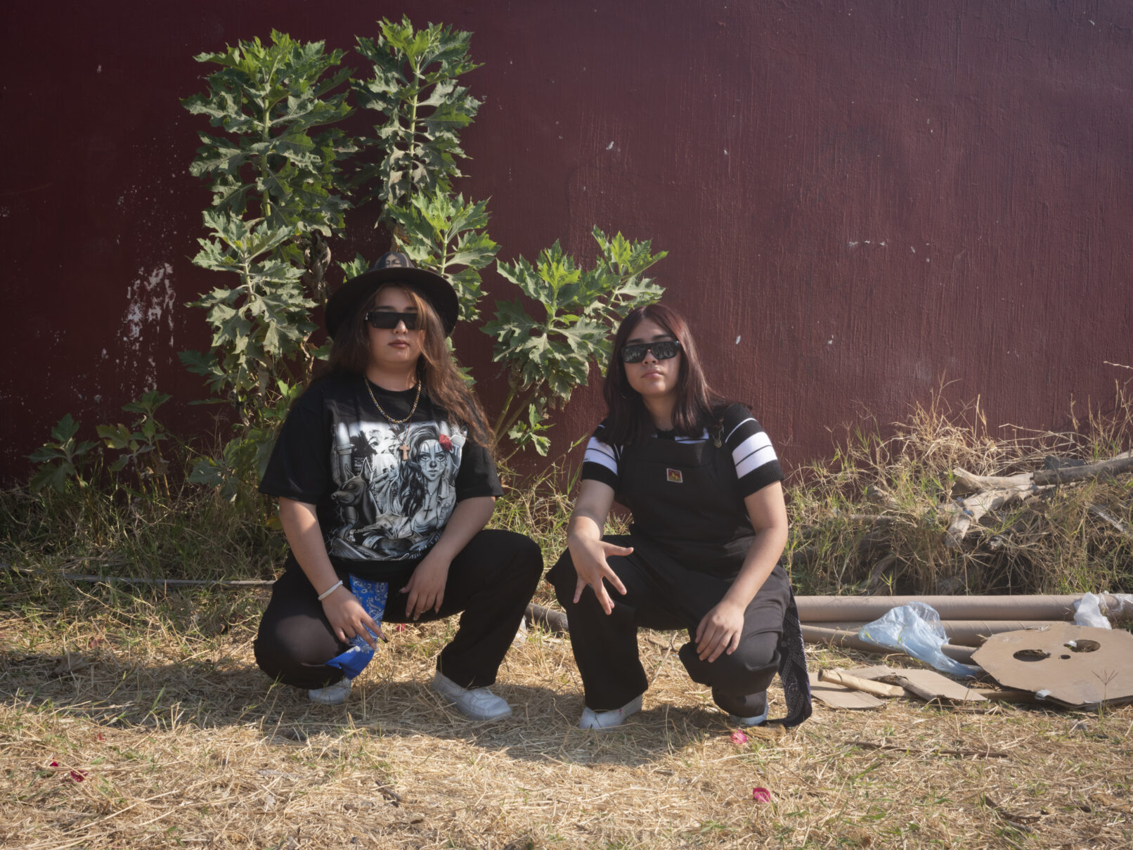 Two young women pose low to the ground in front of a red wall strewn with tall weeds. for the camera, lo