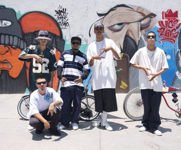 This is a colour photo of 5 young men with deep cholo style standing in front of a graffiti mural. They make gang signs with their hands.