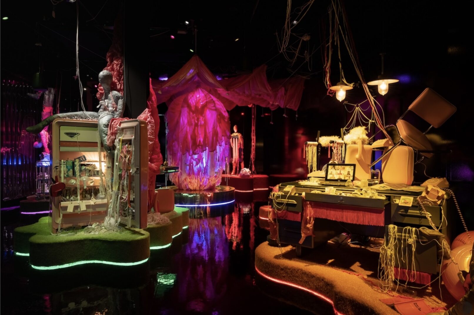 A colour photo of Jess Dobkin's immersive installation called For What It's Worth. This image shows a dark room with warm lighting illuminating three installation stations. One a an open refrigerator strewn with milk and breast pumps, another appears to be a pink, fabric orb, and the last is a work desk strewn chaotically with milk threads, computer, and chairs on top of the desk