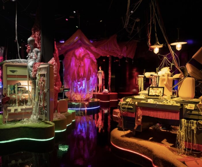 A colour photo of Jess Dobkin's immersive installation called For What It's Worth. This image shows a dark room with warm lighting illuminating three installation stations. One a an open refrigerator strewn with milk and breast pumps, another appears to be a pink, fabric orb, and the last is a work desk strewn chaotically with milk threads, computer, and chairs on top of the desk