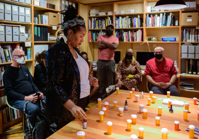 A photo of Jehan Roberson gazing down at a large table arranged with many pill bottles. A small audience of people sit and stand behind her in a library. Jehan is a Black woman. Her hair is tied up with a headband, her large hoop earrings shine. She wears a white shirt and a black blazer.