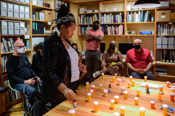 A photo of Jehan Roberson gazing down at a large table arranged with many pill bottles. A small audience of people sit and stand behind her in a library. Jehan is a Black woman. Her hair is tied up with a headband, her large hoop earrings shine. She wears a white shirt and a black blazer.