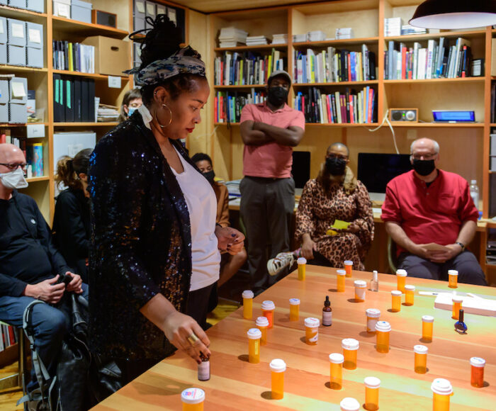 A photo of Jehan Roberson gazing down at a large table arranged with many pill bottles. A small audience of people sit and stand behind her in a library. Jehan is a Black woman. Her hair is tied up with a headband, her large hoop earrings shine. She wears a white shirt and a black blazer.