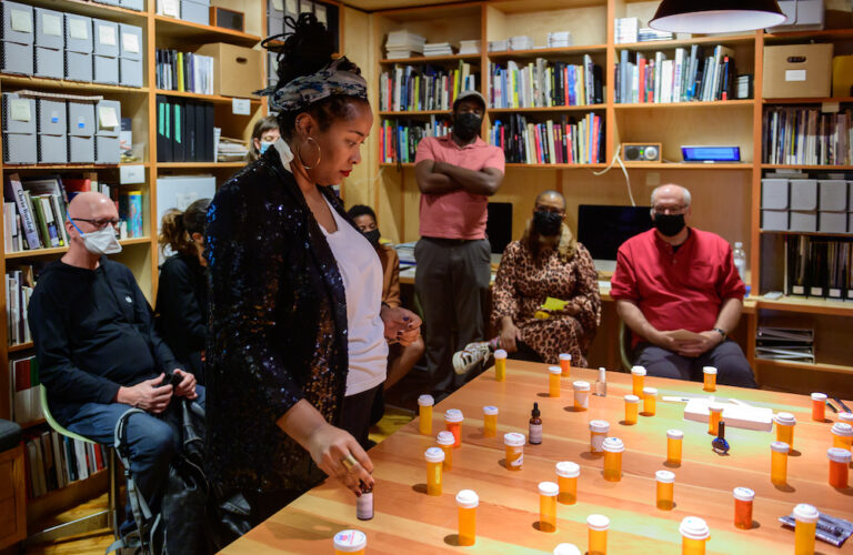 A photo of Jehan Roberson gazing down at a large table arranged with many pill bottles. A small audience of people sit and stand behind her in a library. Jehan is a Black woman. Her hair is tied up with a headband, her large hoop earrings shine. She wears a white shirt and a black blazer.