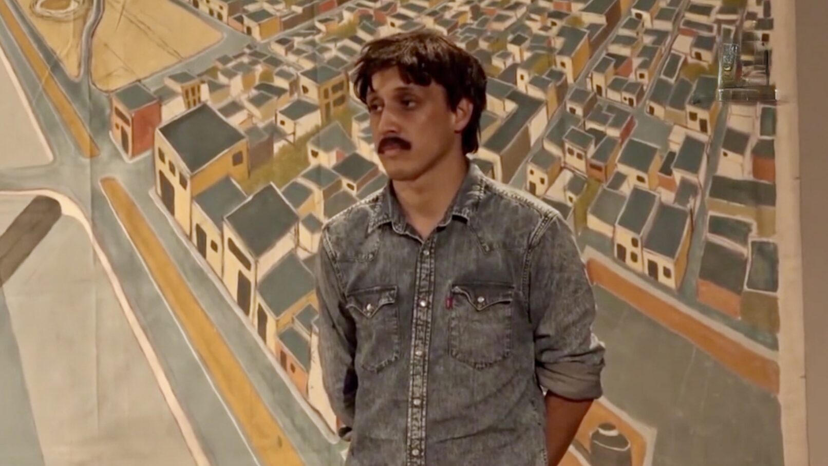 a young man stands in front of a painting of an aerial view of a community of houses.