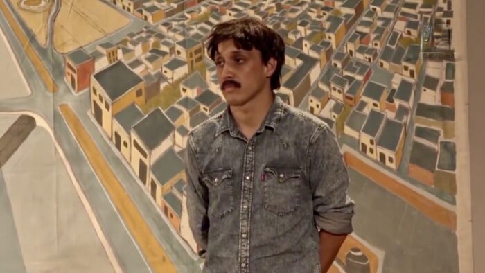 a young man stands in front of a painting of an aerial view of a community of houses.