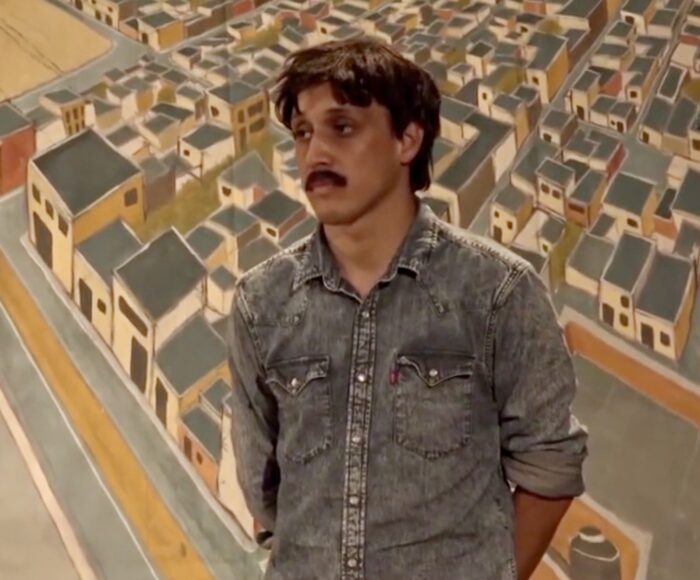 a young man stands in front of a painting of an aerial view of a community of houses.