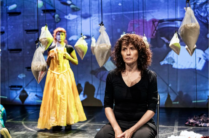 This is a photograph from Teatro Linea de Sombra's production of Amarillo. It pictures a stage with a blue projection. In the background there are many large bags filled with sand suspended around the set. In the foreground, a woman with dark curly hair wearing black clothing sits in a chair. In the background, a person stands wearing a long, flowy, yellow garment.
