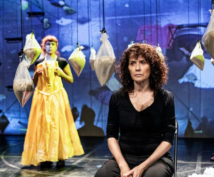 This is a photograph from Teatro Linea de Sombra's production of Amarillo. It pictures a stage with a blue projection. In the background there are many large bags filled with sand suspended around the set. In the foreground, a woman with dark curly hair wearing black clothing sits in a chair. In the background, a person stands wearing a long, flowy, yellow garment.