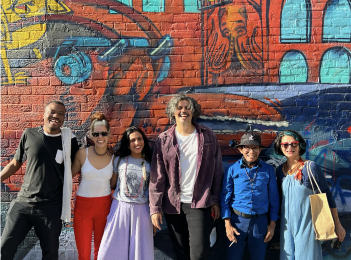 A group of six smiling people gathering for a picture in front of a big, colourful graffiti wall.