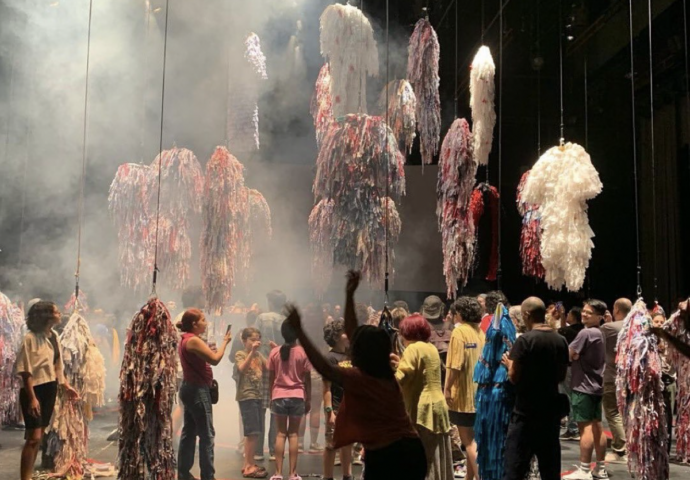 This photograph pictures a stage with dozens of colourful, shaggy costumes suspended in the air. Below them is a crowd of people gathered, looking at and taking pictures of the costumes. There's fog in the air against the black background of the theatre.