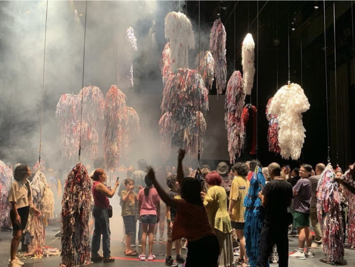 This photograph pictures a stage with dozens of colourful, shaggy costumes suspended in the air. Below them is a crowd of people gathered, looking at and taking pictures of the costumes. There's fog in the air against the black background of the theatre.