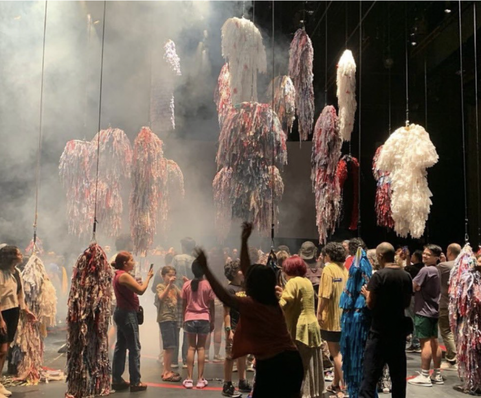 This photograph pictures a stage with dozens of colourful, shaggy costumes suspended in the air. Below them is a crowd of people gathered, looking at and taking pictures of the costumes. There's fog in the air against the black background of the theatre.
