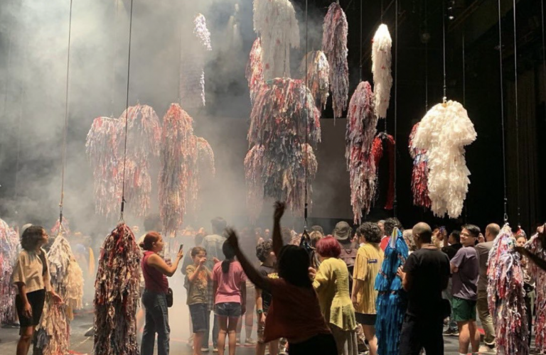 This photograph pictures a stage with dozens of colourful, shaggy costumes suspended in the air. Below them is a crowd of people gathered, looking at and taking pictures of the costumes. There's fog in the air against the black background of the theatre.